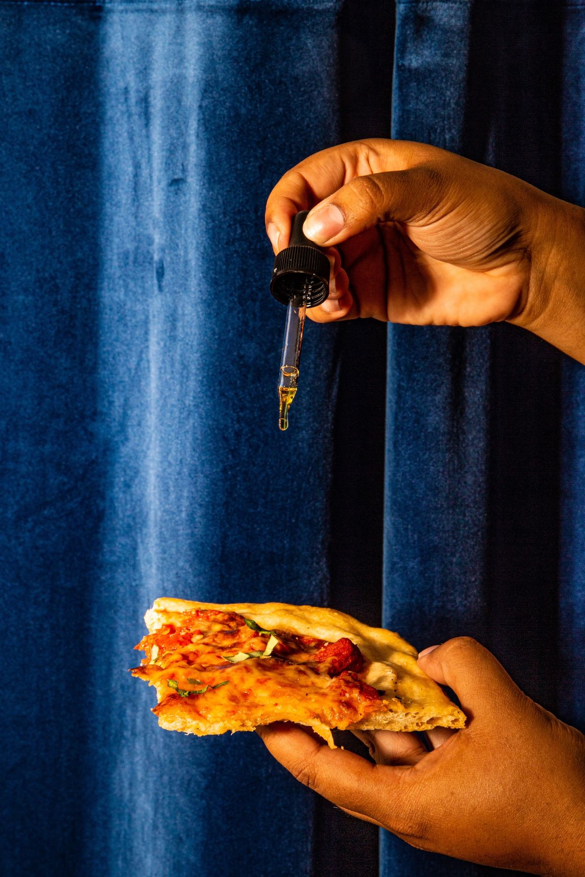 A hand adding a drop of tincture to a slice of pizza against a blue velvet curtain