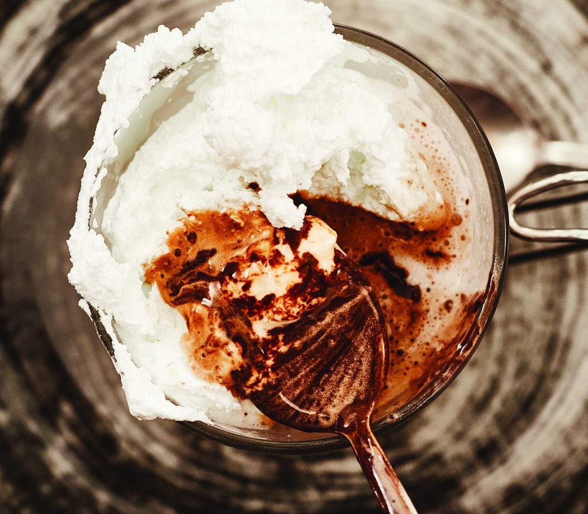 A spoon breaking through whipped cream into a glass of hot chocolate, seen from above